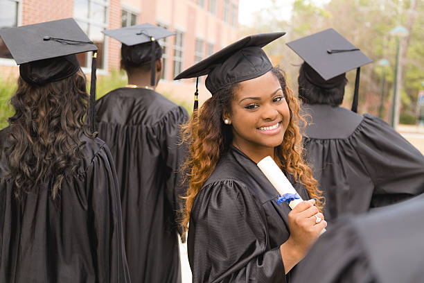 Candid moment of African descent female excitedly holding her diploma after the college graduation ceremony.  Her friends around her.  School building background on campus.