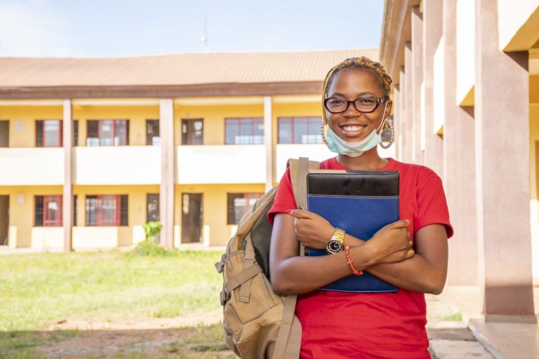 A young African female student with a facemask holding her textbooks at a campus area